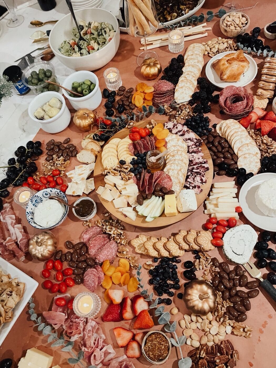 a giant cheese board spread across a dining room table on brown butcher paper with cheeses, salami, crackers, fruit, nuts, candles and gold pumpkins