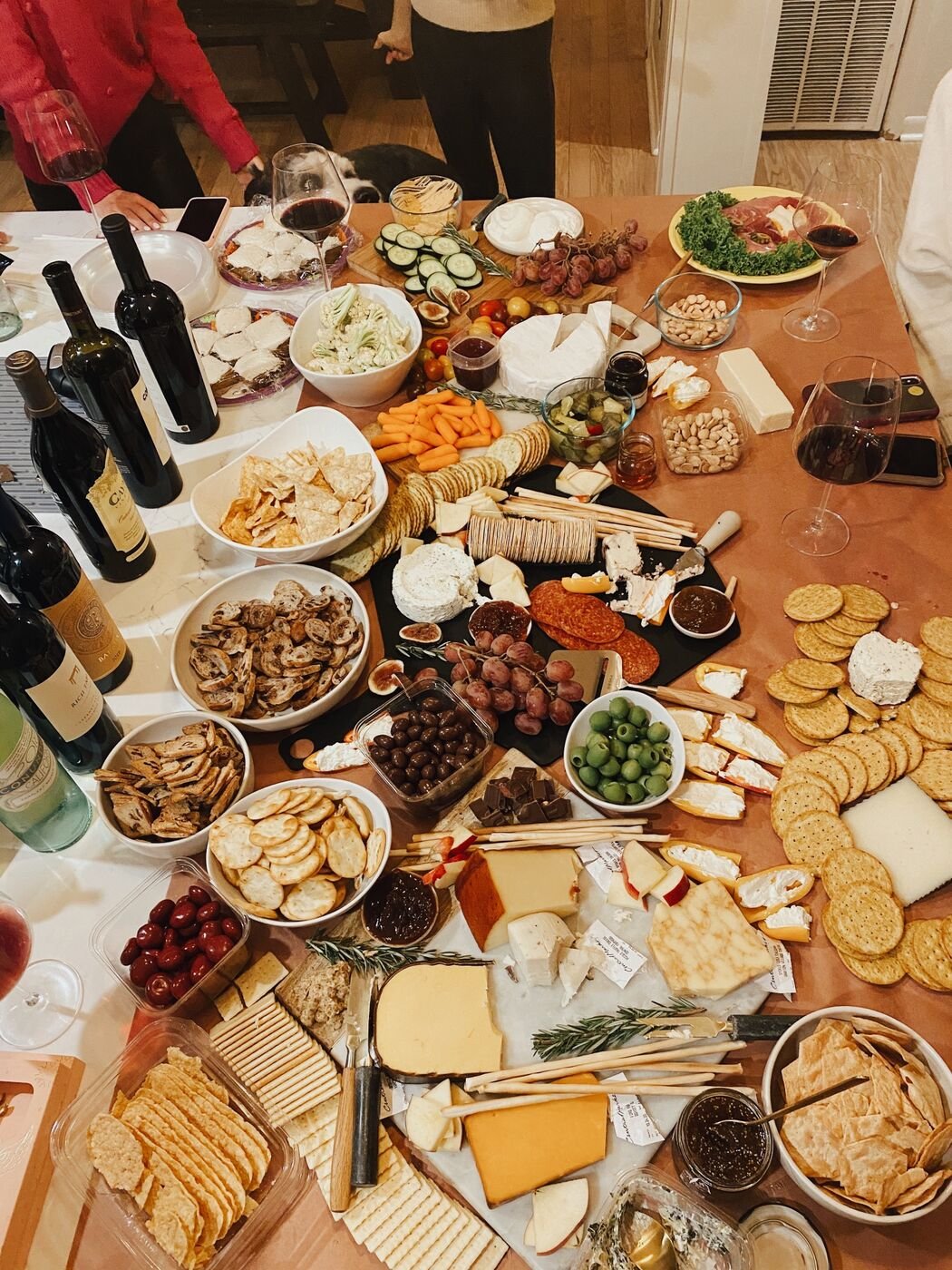 a dining room table covered in cheeses, crackers, olives, jams and bottles of red wine at cheesegiving